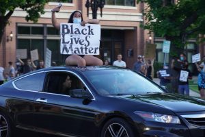 A protestor holding a "Black Lives Matter" poster sits on top of a car as it drives down Main Street in Logan, UT.