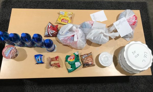 Photograph of multiple bottles of water, pre-packaged cookies, chips, and snacks. There are also covered containers of hot food.