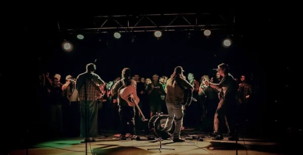 Four members of a band look into a crowd in low light. They carry a mix of instruments including a banjo and a an acoustic guitar.