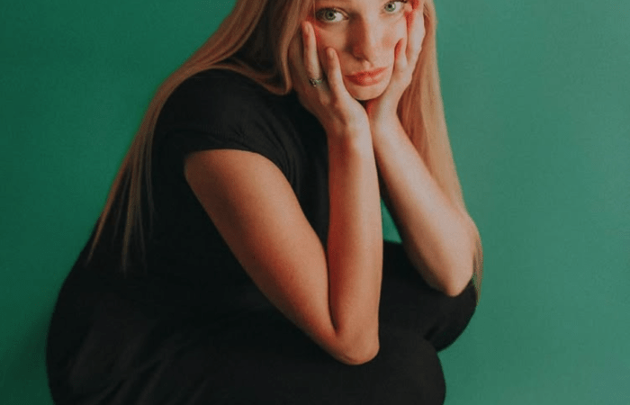 A woman with blonde hair squats in front of a teal background. She has her hands on her face, elbows on her knees, and is looking at the camera. She wears an all black outfit.